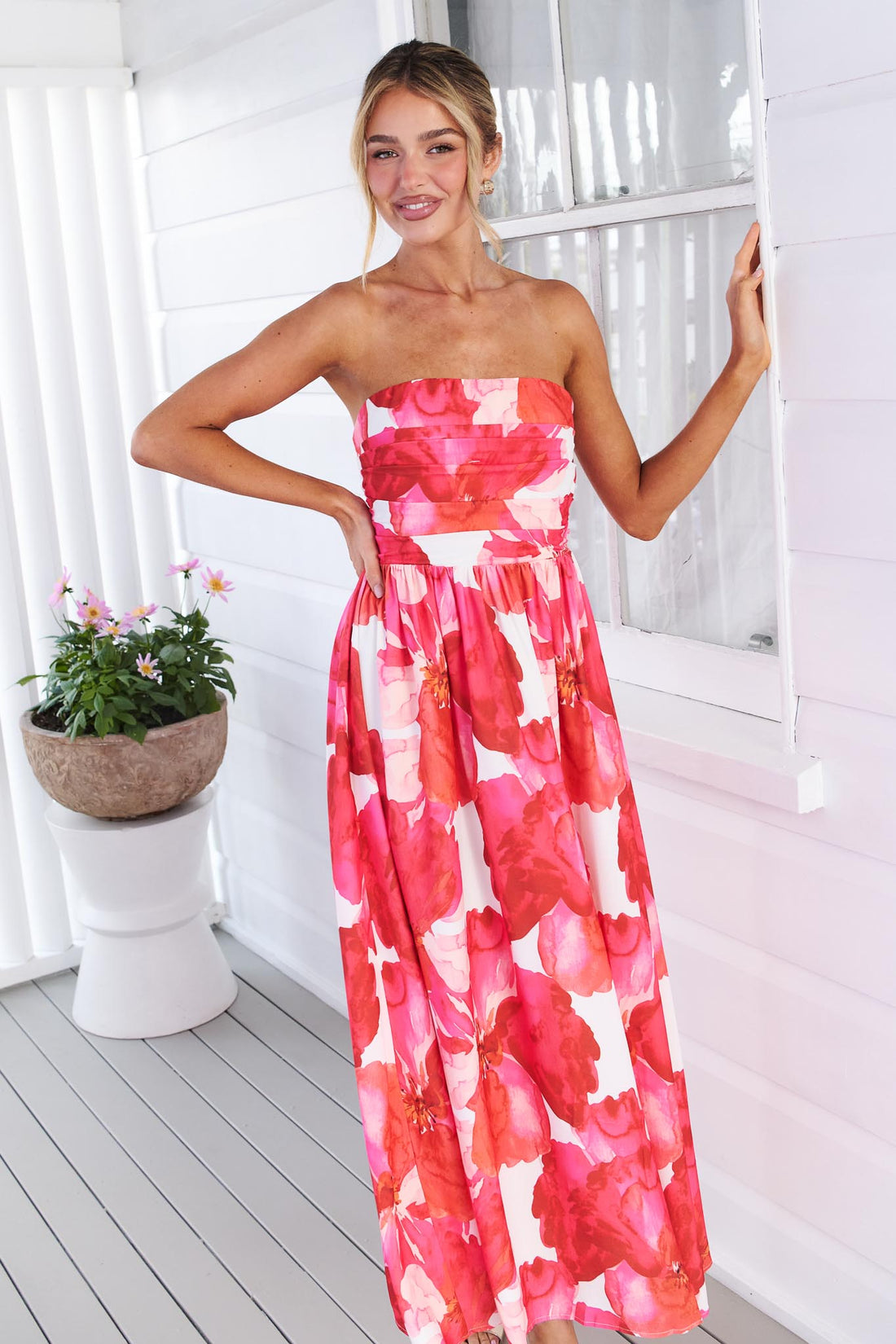 Back view of a woman wearing the minnie + MAXXIE Sienna Strapless pink floral event dress, standing against a white panelled wall with potted flowers in the background.