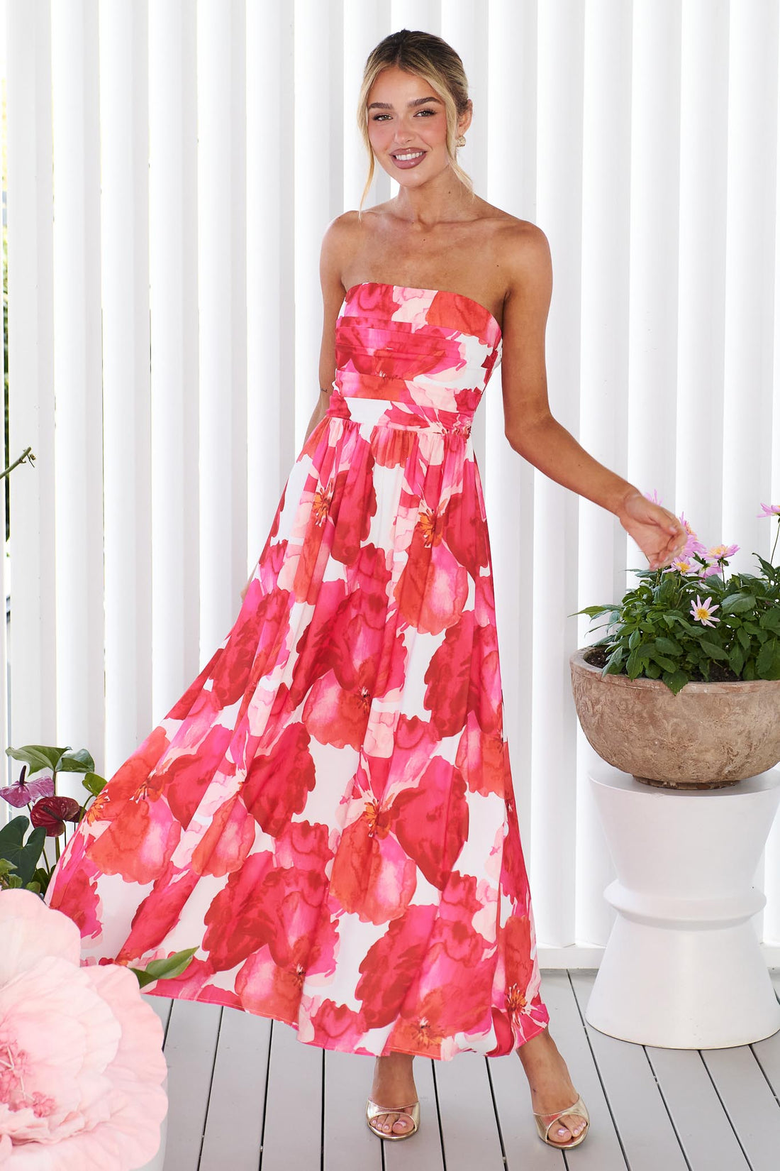 A woman wearing the minnie + MAXXIE Sienna Strapless pink floral event dress, standing against a white panelled wall with potted flowers in the background.