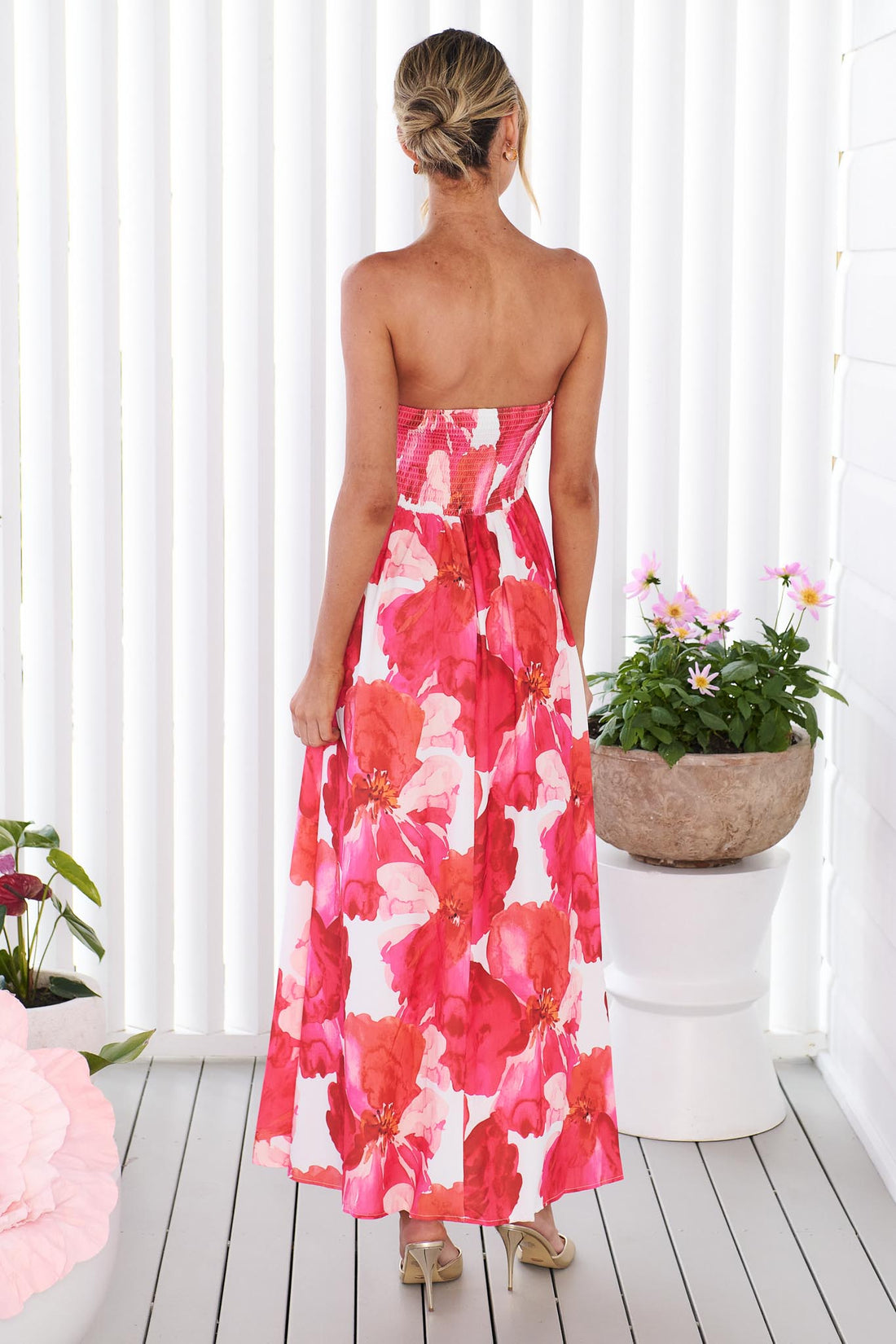 Back view of a woman wearing the minnie + MAXXIE Sienna Strapless pink floral event dress, standing against a white panelled wall with potted flowers in the background.