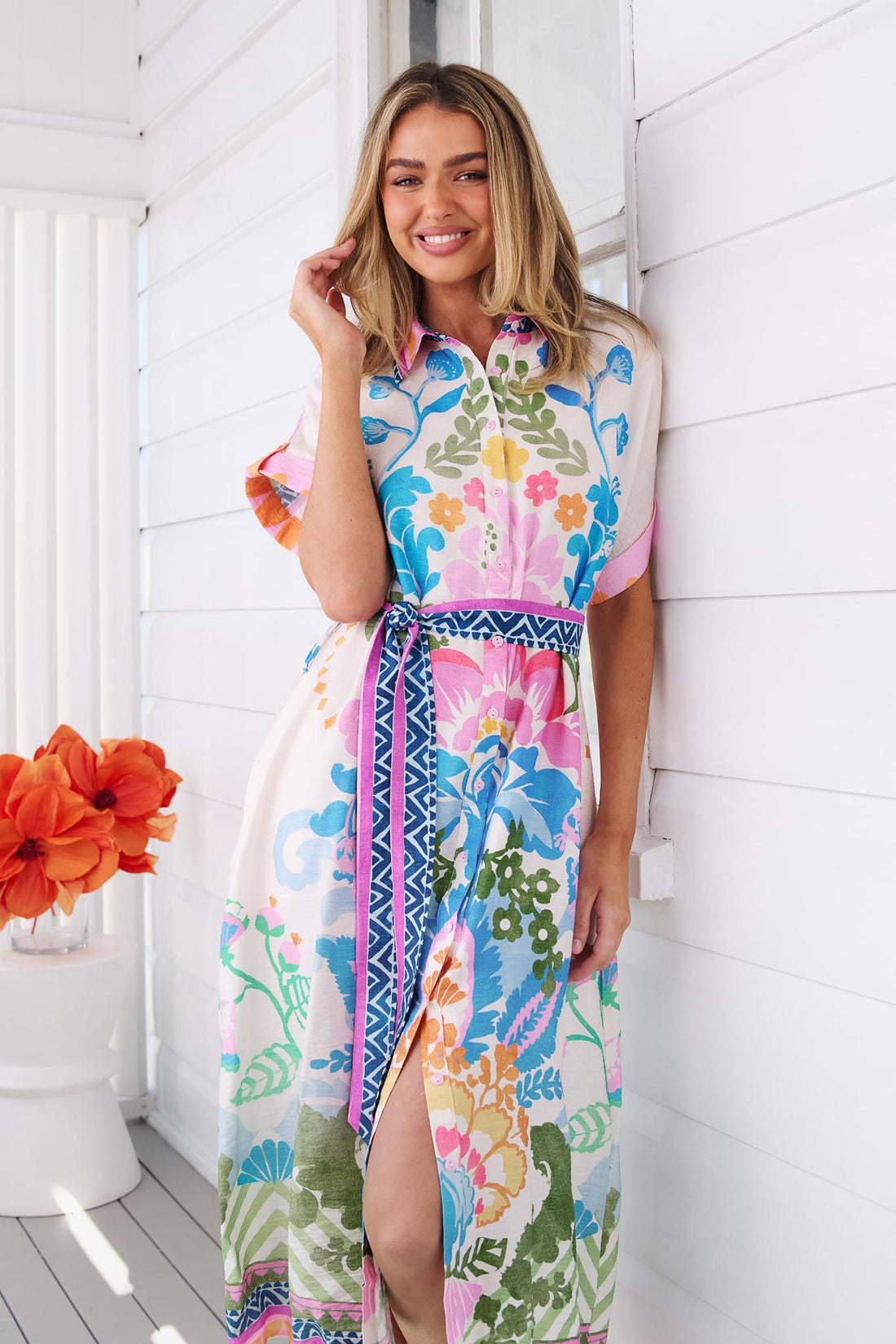 A woman wearing the colourful, short-sleeved minnie + MAXXIE Solana midi dress with a vibrant floral and geometric print, standing on a white wooden porch.
