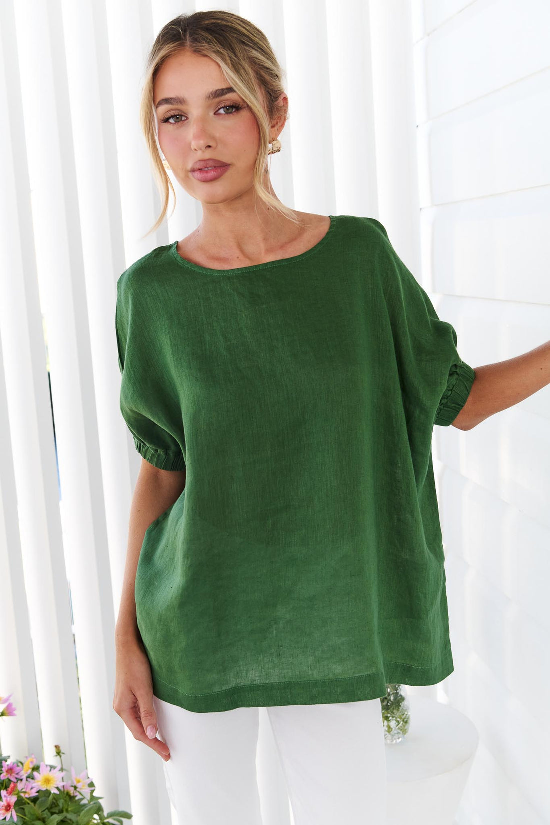 A woman wearing the minnie + MAXXIE studio linen top in emerald green with white jeans, standing in front of a white panelled wall