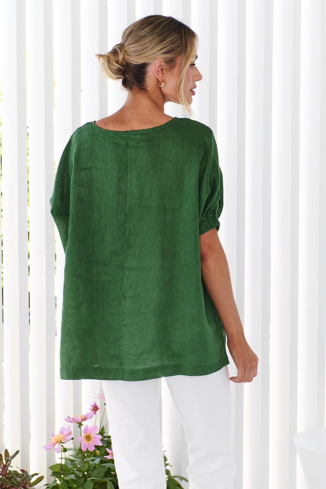 Back view of a woman wearing the minnie + MAXXIE studio linen top in emerald green with white jeans, standing in front of a white panelled wall