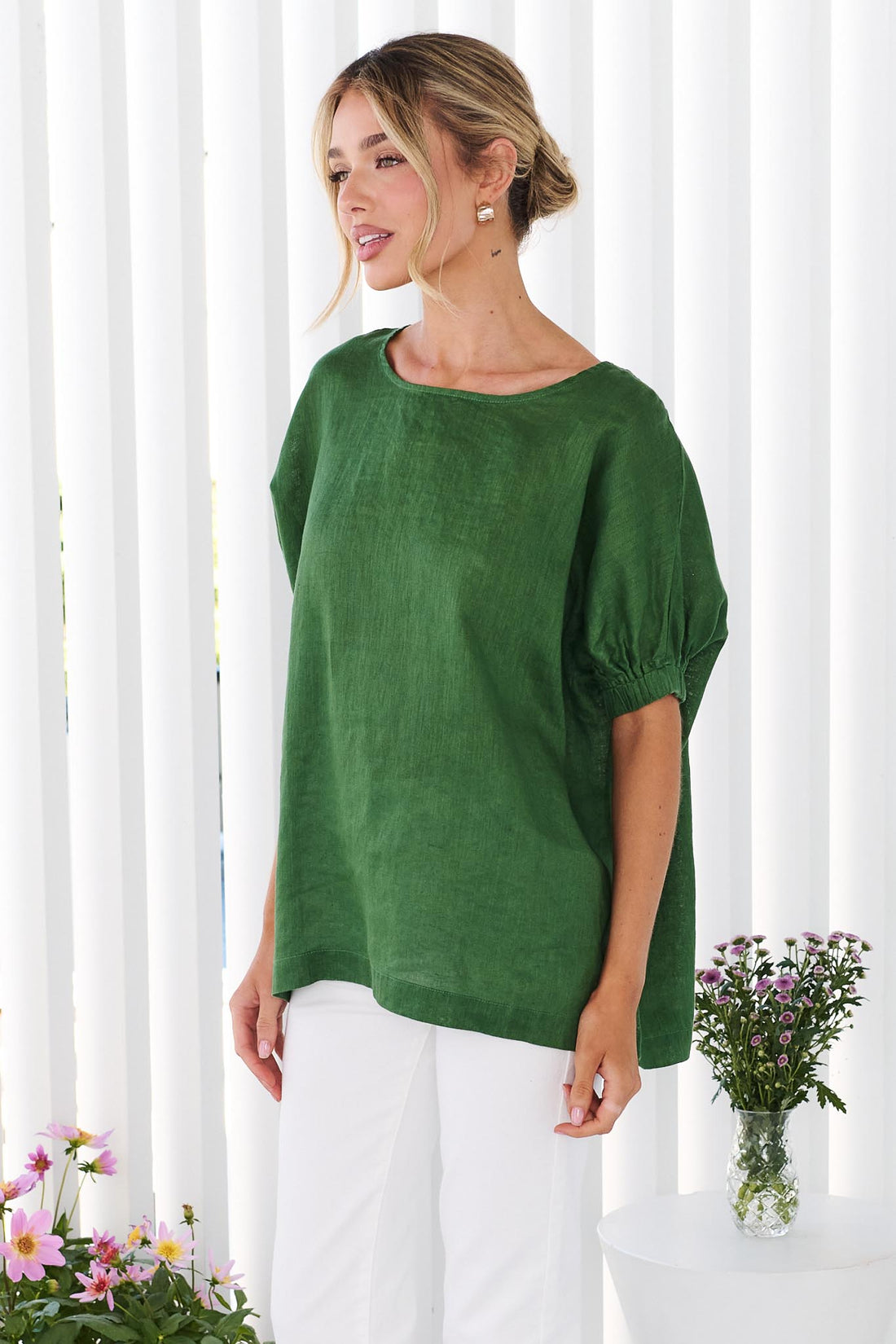 Side view of a woman wearing the minnie + MAXXIE studio linen top in emerald green with white jeans, standing in front of a white panelled wall