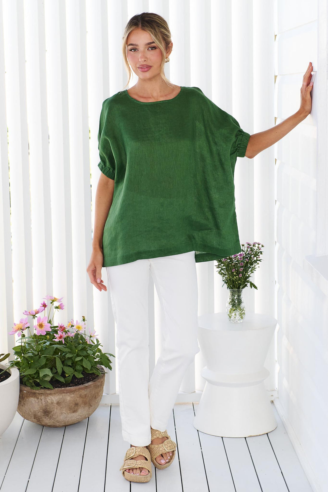 A woman wearing the minnie + MAXXIE studio linen top in emerald green with white jeans, standing in front of a white panelled wall