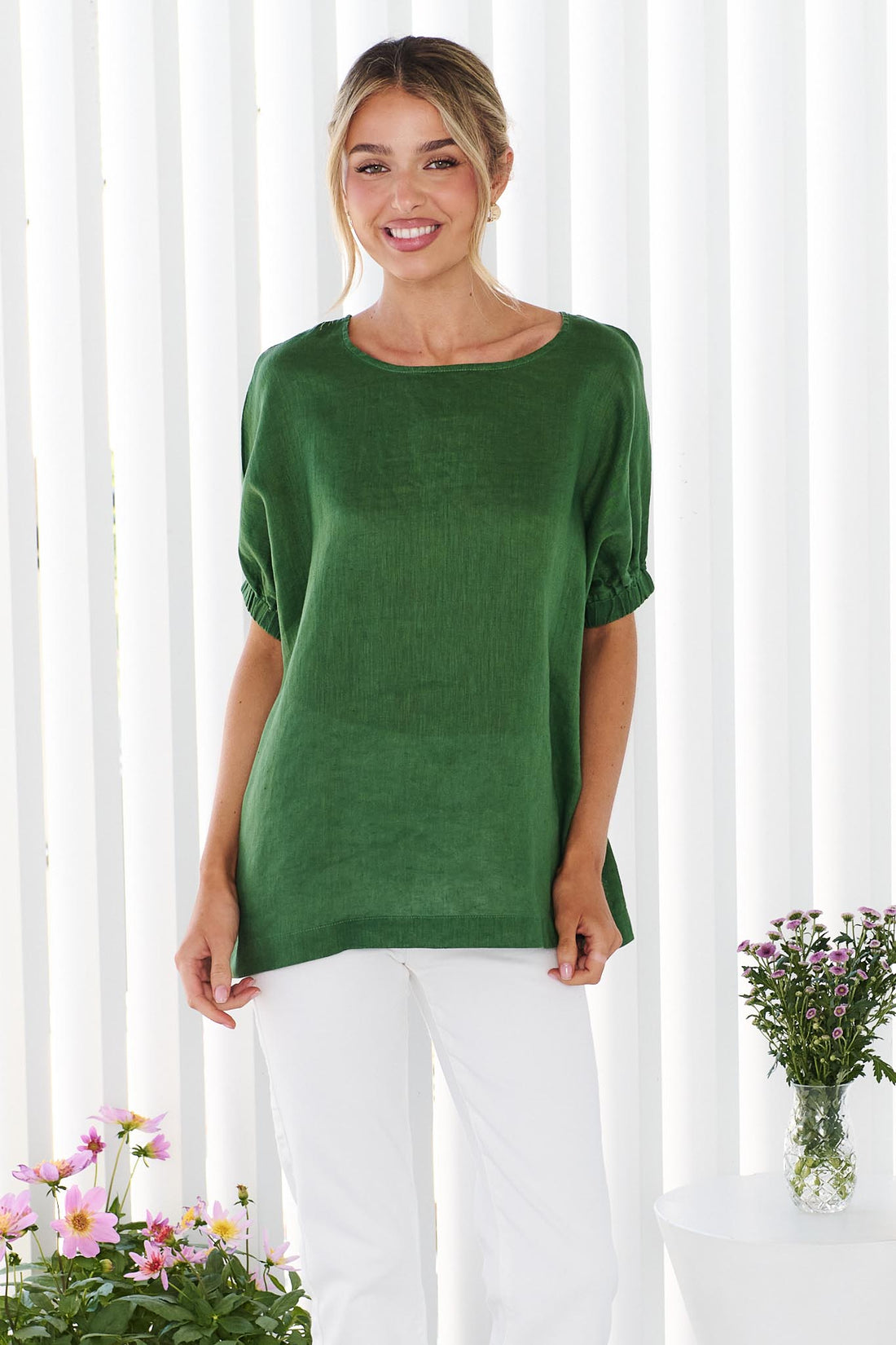 A woman wearing the minnie + MAXXIE studio linen top in emerald green with white jeans, standing in front of a white panelled wall