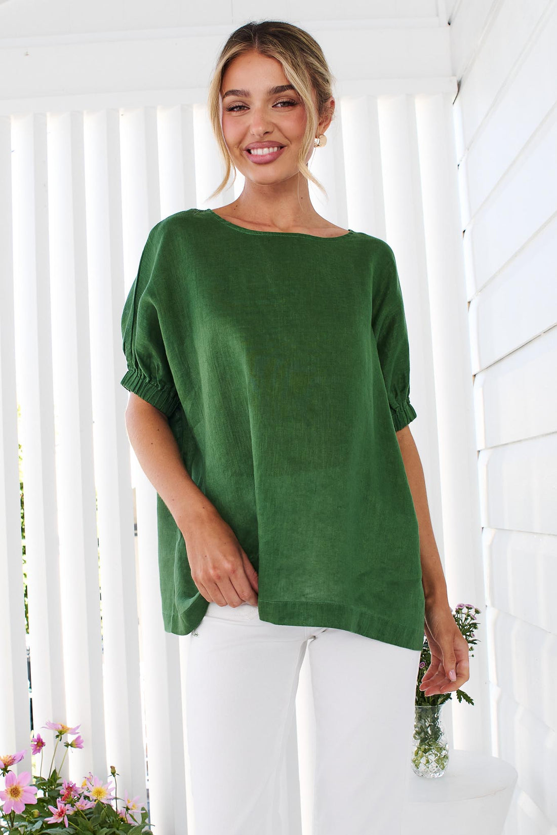 A woman wearing the minnie + MAXXIE studio linen top in emerald green with white jeans, standing in front of a white panelled wall