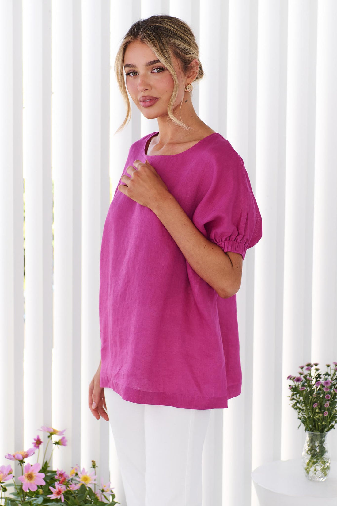 Side view of a woman wearing the minnie + MAXXIE studio linen top in Magenta pink standing in front of a white panelled wall
