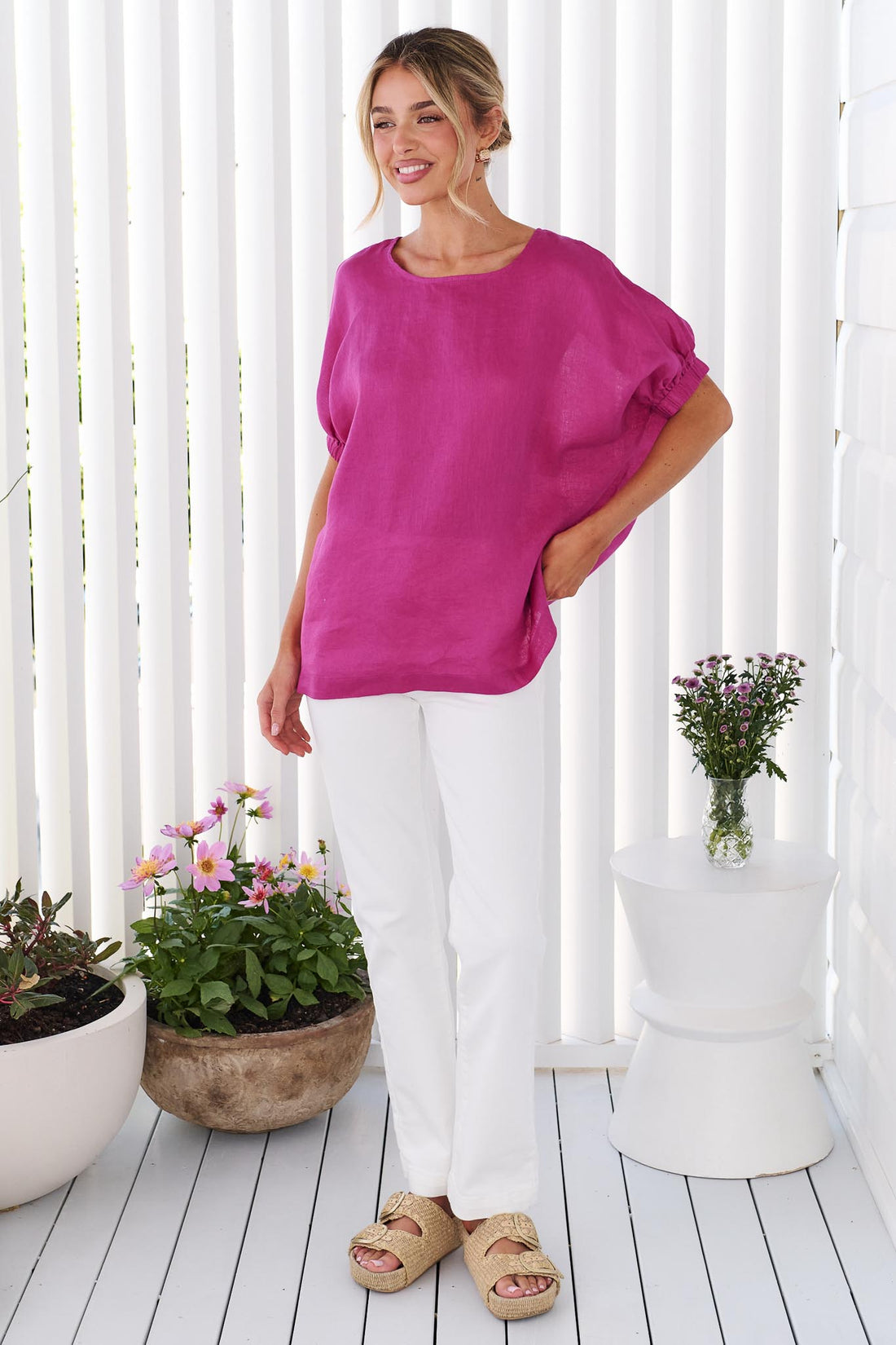 A woman wearing the minnie + MAXXIE studio linen top in Magenta pink with white jeans, standing in front of a white panelled wall
