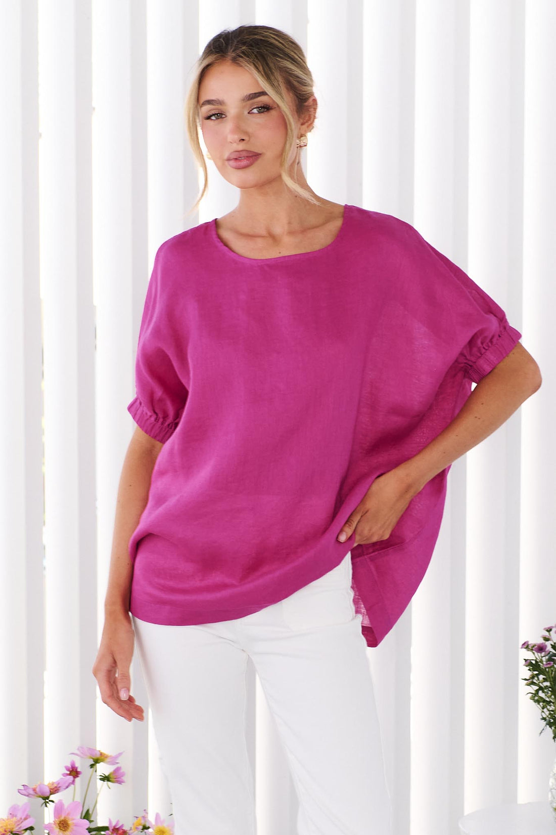 A woman wearing the minnie + MAXXIE studio linen top in Magenta pink with white jeans, standing in front of a white panelled wall
