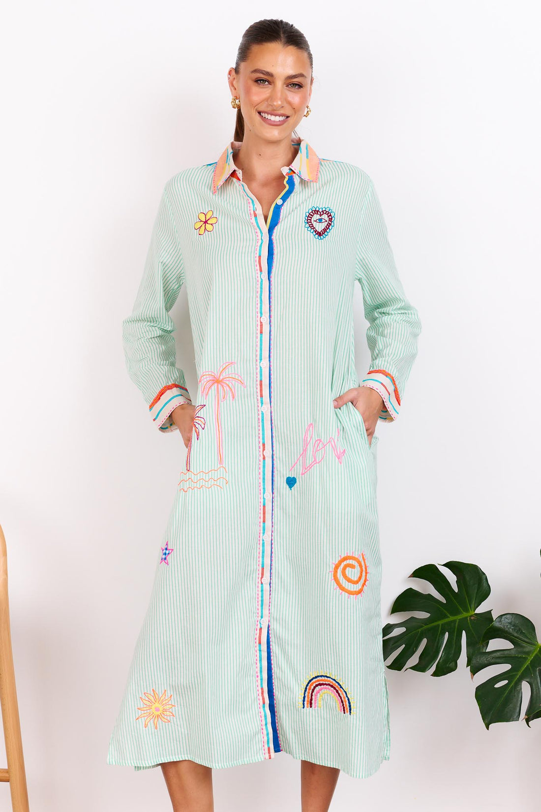 Woman wearing the minnie + MAXXIE Summer Love Cotton shirt dress with a green pinstripe print and colourful embroidery details, standing against a white wall.