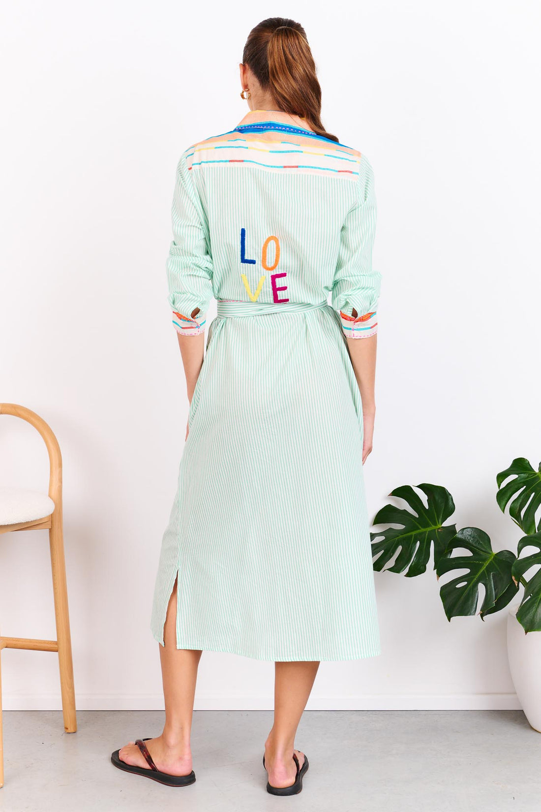 Back view of a woman wearing the minnie + MAXXIE Summer Love Cotton shirt dress with a green pinstripe print, colourful embroidery details and a matching waist tie; standing against a white wall.