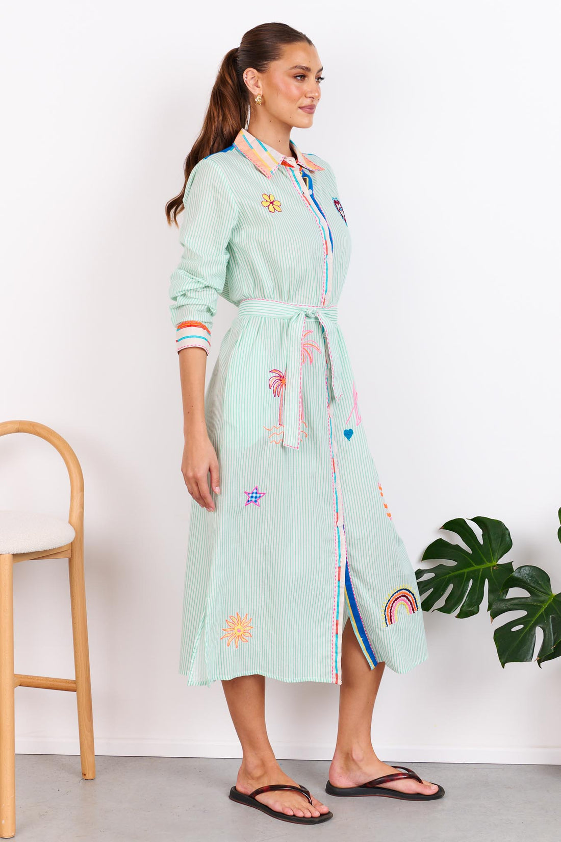 Woman wearing the minnie + MAXXIE Summer Love Cotton shirt dress with a green pinstripe print, colourful embroidery details and a matching waist tie; standing against a white wall.