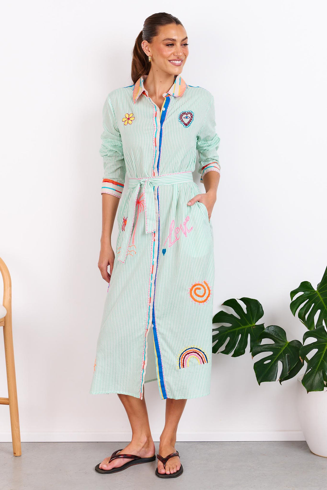 Woman wearing the minnie + MAXXIE Summer Love Cotton shirt dress with a green pinstripe print, colourful embroidery details and a matching waist tie; standing against a white wall.