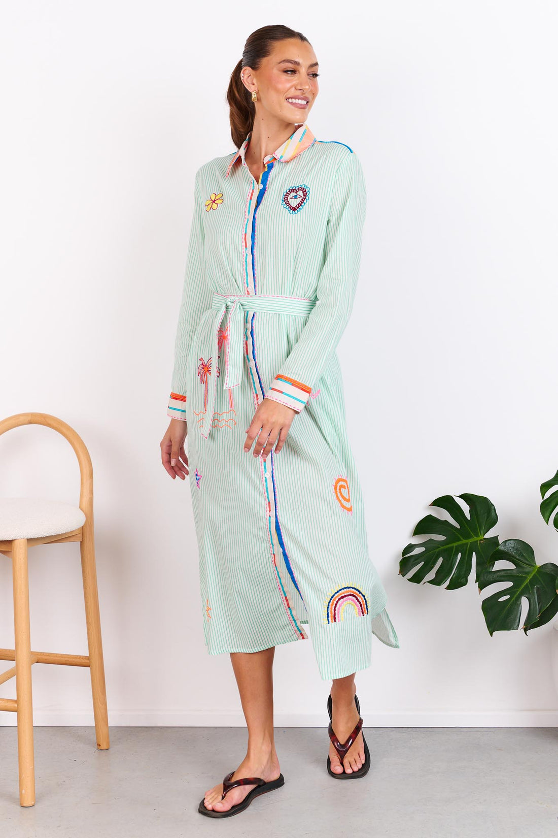 Woman wearing the minnie + MAXXIE Summer Love Cotton shirt dress with a green pinstripe print, colourful embroidery details and a matching waist tie; standing against a white wall.