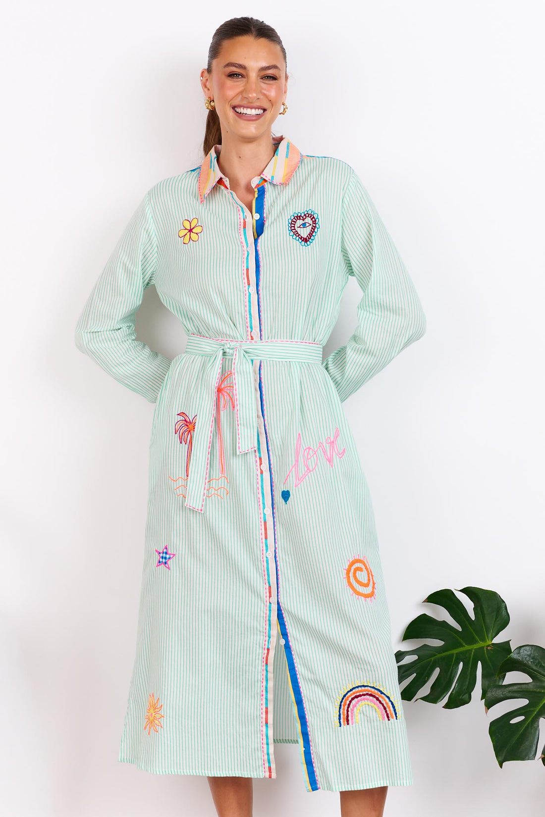Woman wearing the minnie + MAXXIE Summer Love Cotton shirt dress with a green pinstripe print, colourful embroidery details and a matching waist tie; standing against a white wall.