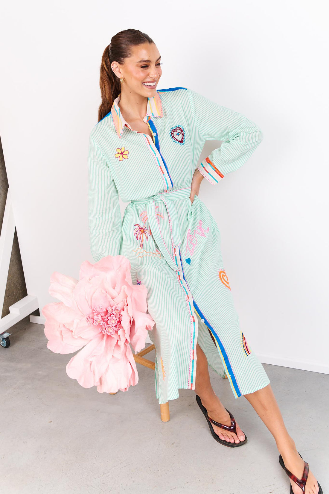 Woman wearing the minnie + MAXXIE Summer Love Cotton shirt dress with a green pinstripe print, colourful embroidery details and a matching waist tie; sitting on a stool against a white wall.