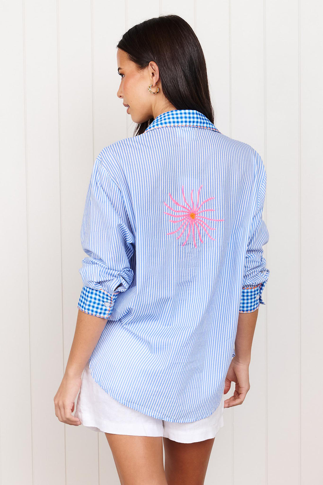 Back view of a woman wearing the minnie + MAXXIE Tutti Cotton Shirt, a light blue and white striped shirt with gingham trims, featuring neon pink and orange embroidered sun.