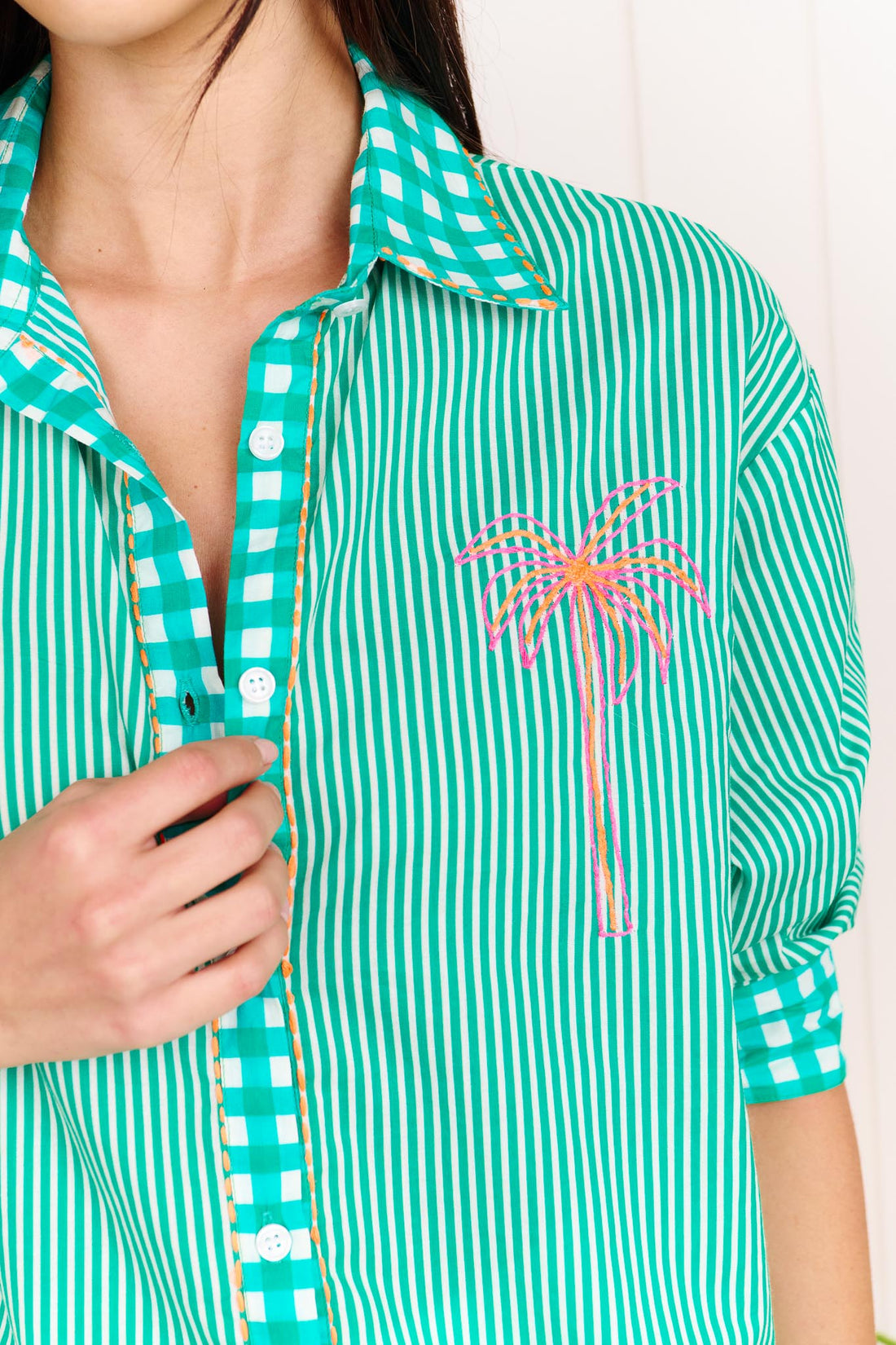 Close up of a woman wearing the minnie + MAXXIE Tutti Cotton Shirt, a bright green and white striped shirt with gingham trims, featuring neon pink and orange palm tree, heart and shell embroidery.