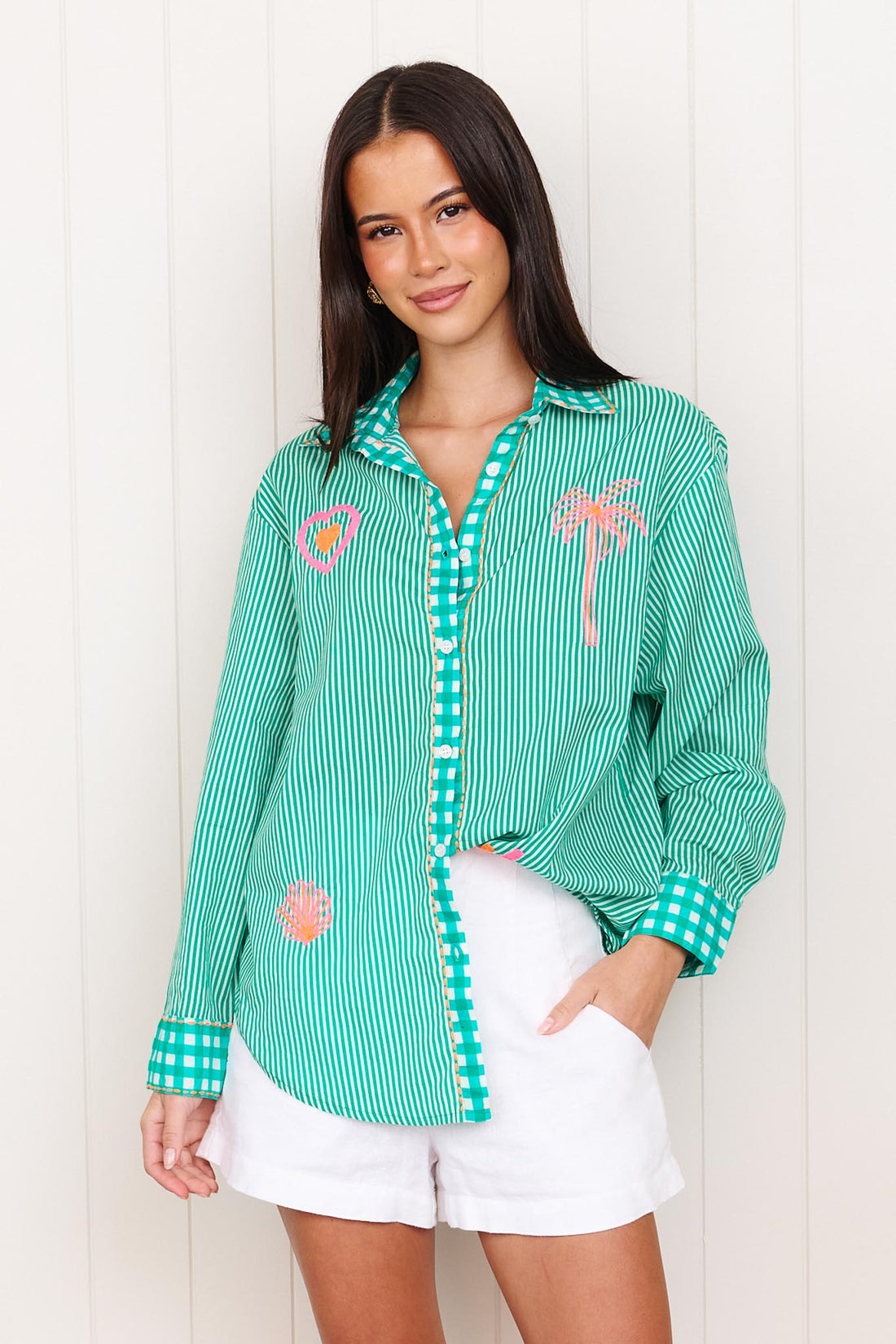 A woman wearing the minnie + MAXXIE Tutti Cotton Shirt, a bright green and white striped shirt with gingham trims, featuring neon pink and orange palm tree, heart and shell embroidery.