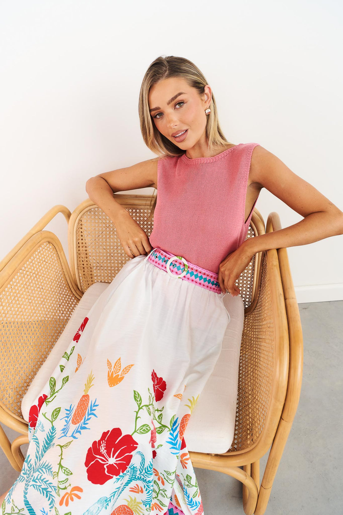 Woman sitting on a wicker chair wearing the Valencia floral maxi skirt with pineapple print and matching belt styled with pink Villa knit tank