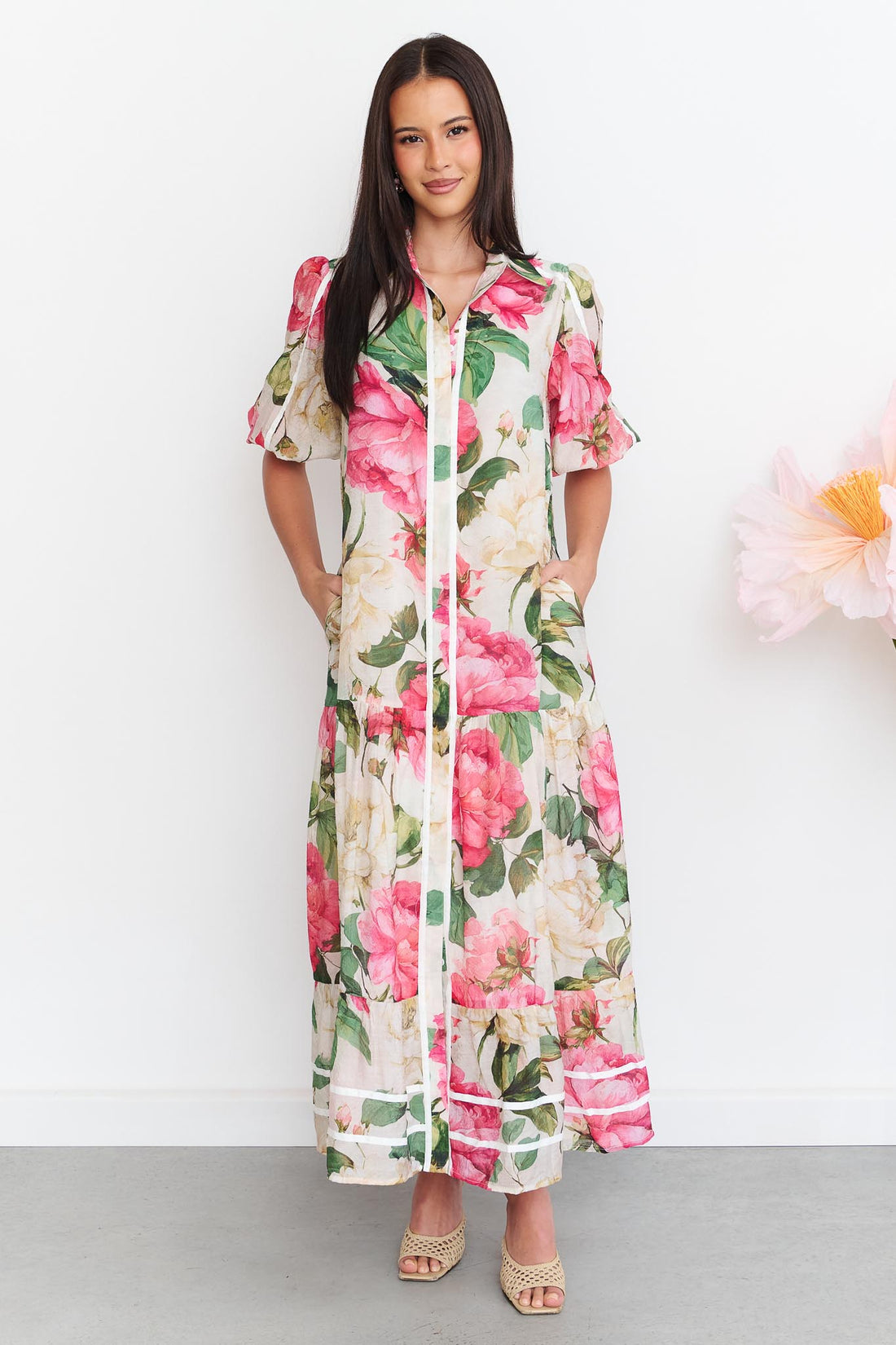 A woman wearing the minnie + MAXXIE Valois maxi shirt dress with a vibrant pink and green floral print. The dress features contrast white ribbon trims, and short puff sleeves.
