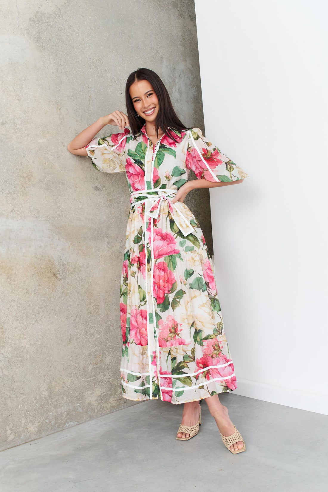 A woman wearing the minnie + MAXXIE Valois maxi shirt dress with a vibrant pink and green floral print. The dress features a matching fabric waist tie, and short puff sleeves.