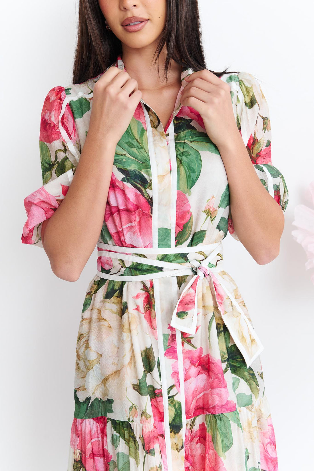 Detail of a woman wearing the minnie + MAXXIE Valois maxi shirt dress with a vibrant pink and green floral print. The dress features a matching fabric waist tie, and short puff sleeves.