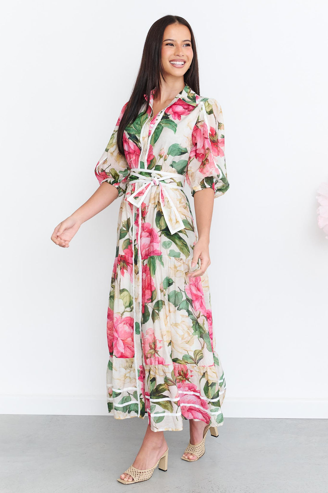 A woman wearing the minnie + MAXXIE Valois maxi shirt dress with a vibrant pink and green floral print. The dress features a matching fabric waist tie, and short puff sleeves.