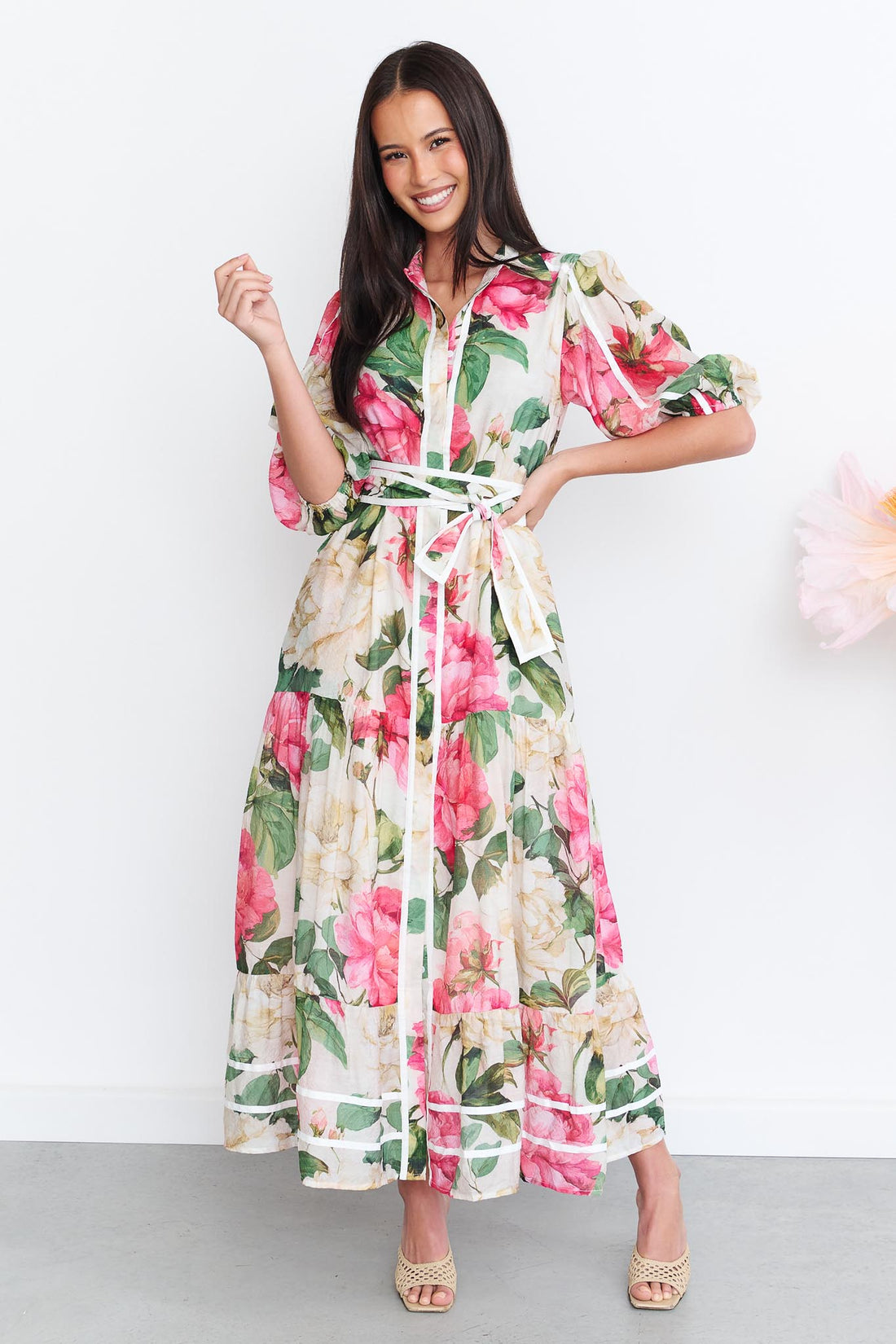 A woman wearing the minnie + MAXXIE Valois maxi shirt dress with a vibrant pink and green floral print. The dress features a matching fabric waist tie, and short puff sleeves.