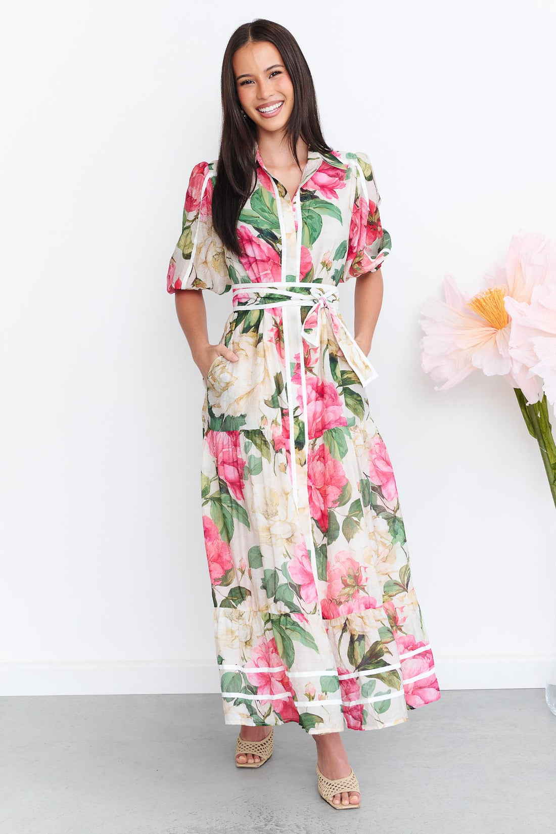 A woman wearing the minnie + MAXXIE Valois maxi shirt dress with a vibrant pink and green floral print. The dress features a matching fabric waist tie, and short puff sleeves.