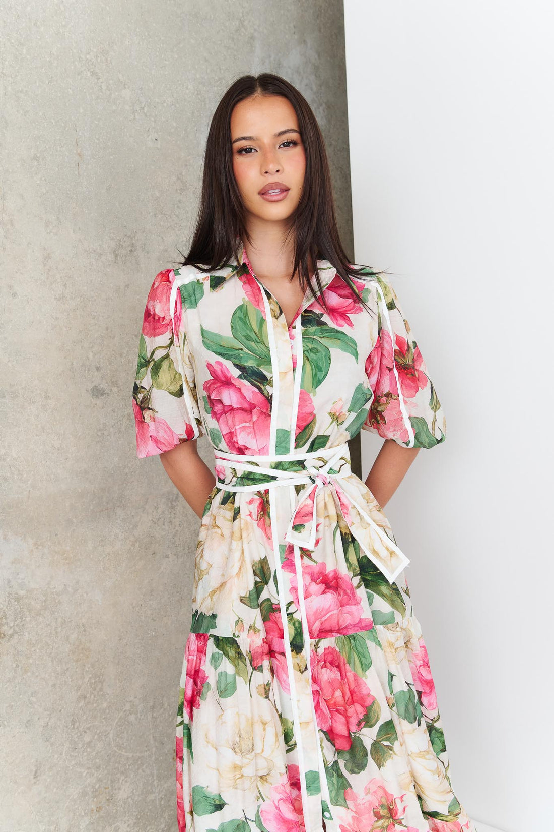 A woman wearing the minnie + MAXXIE Valois maxi shirt dress with a vibrant pink and green floral print. The dress features a matching fabric waist tie, and short puff sleeves.