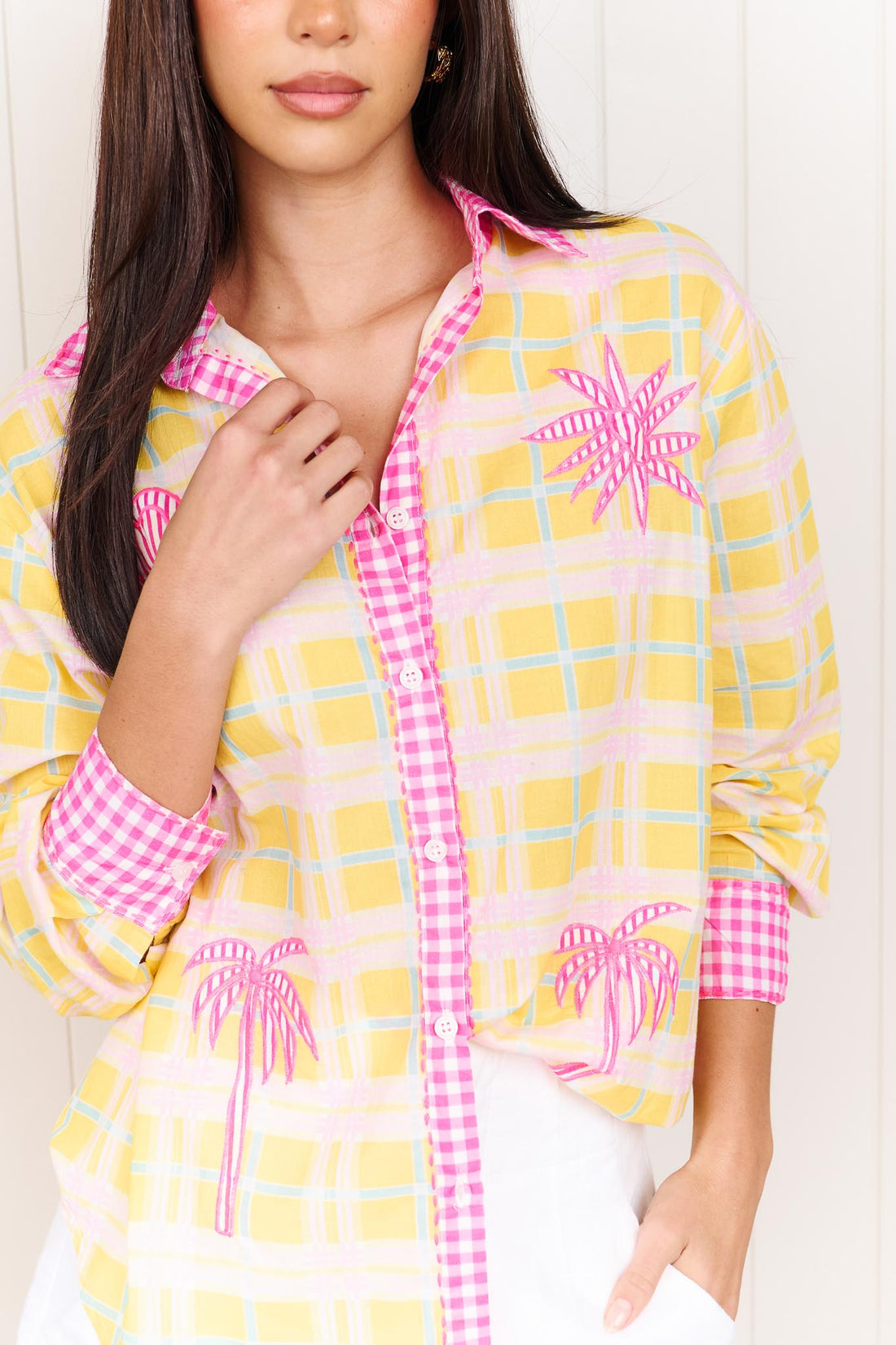 Detail of awoman wearing the minnie + MAXXIE Verano Cotton Shirt in Yellow checks with pink gingham trims and palm embroidery, standing against a white wall.