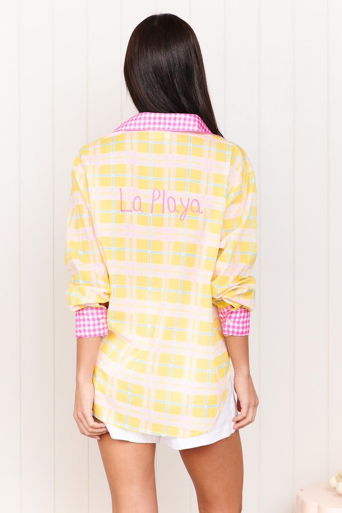 Back view of a woman wearing the minnie + MAXXIE Verano Cotton Shirt in Yellow checks with pink gingham trims and La Playa embroidery, standing against a white wall.