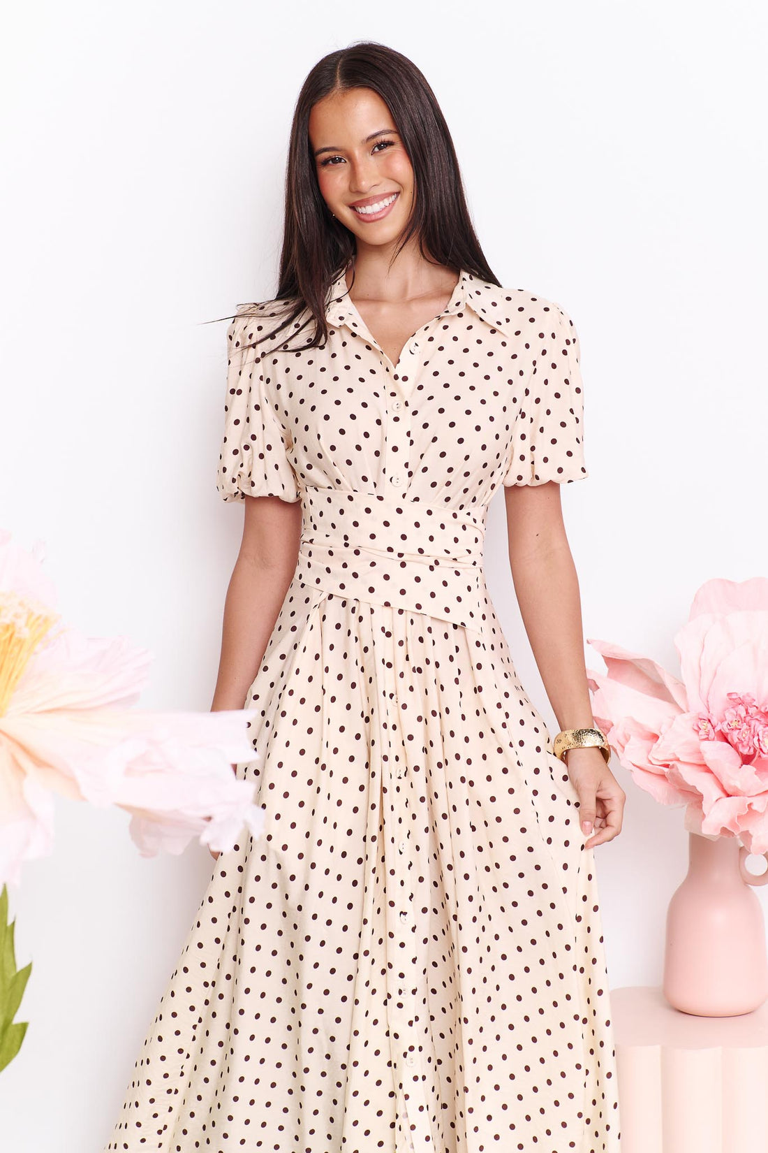 A model wearing the minnie + MAXXIE Viella Midi Dress in buttermilk with brown polka dots and a crossover waist, standing in front of a white wall.
