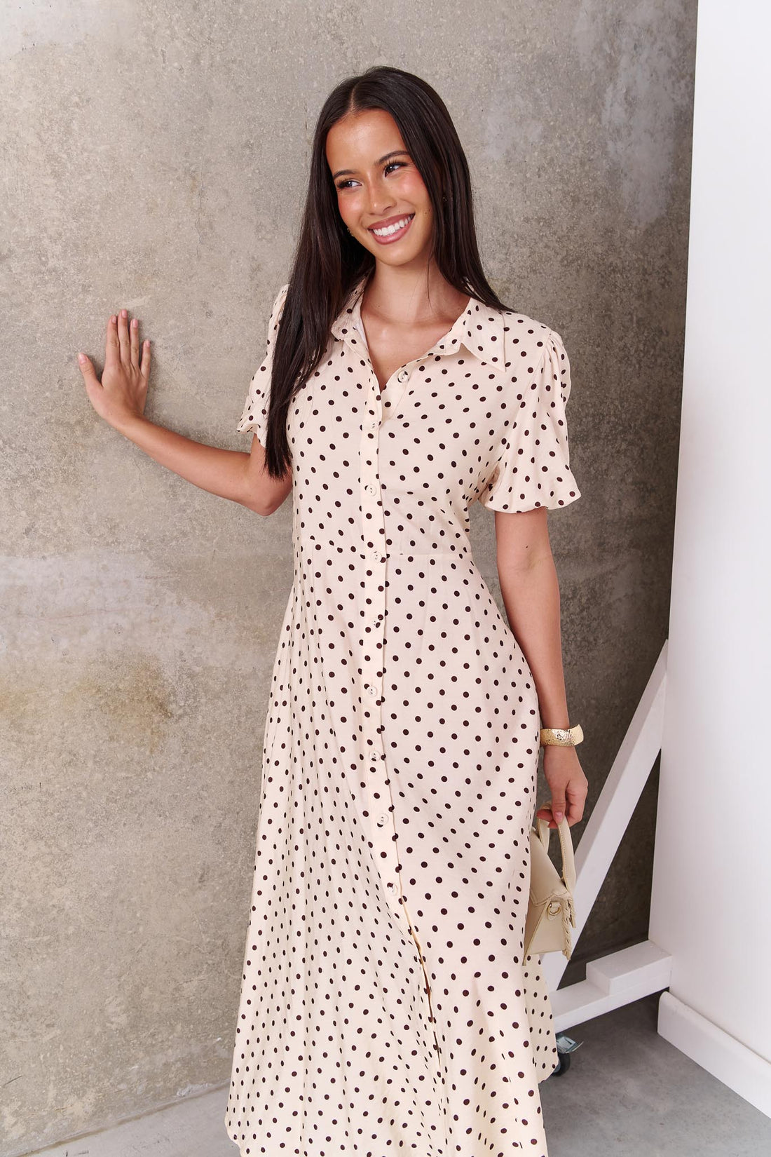 A model wearing the minnie + MAXXIE Viella Midi Dress in buttermilk with brown polka dots and button front, standing in front of a concrete wall.