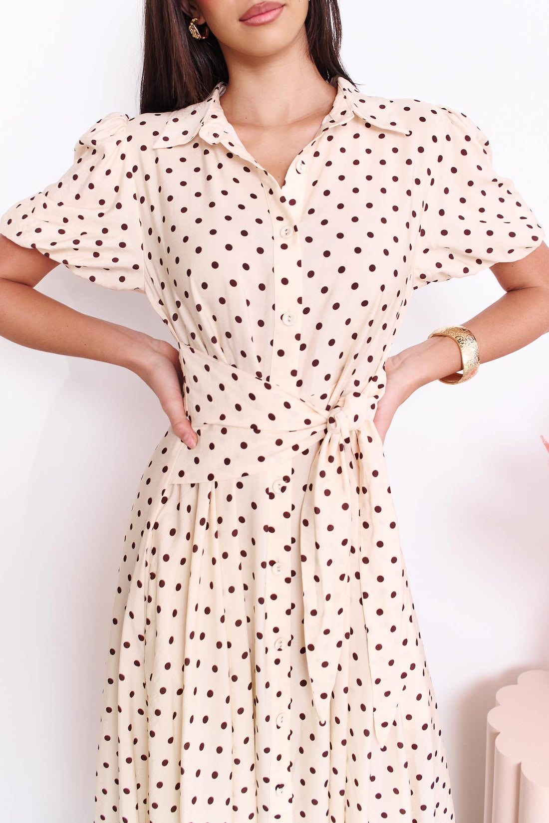 Close up of a model wearing the minnie + MAXXIE Viella Midi Dress in buttermilk with brown polka dots and a front waist tie, standing in front of a white wall.