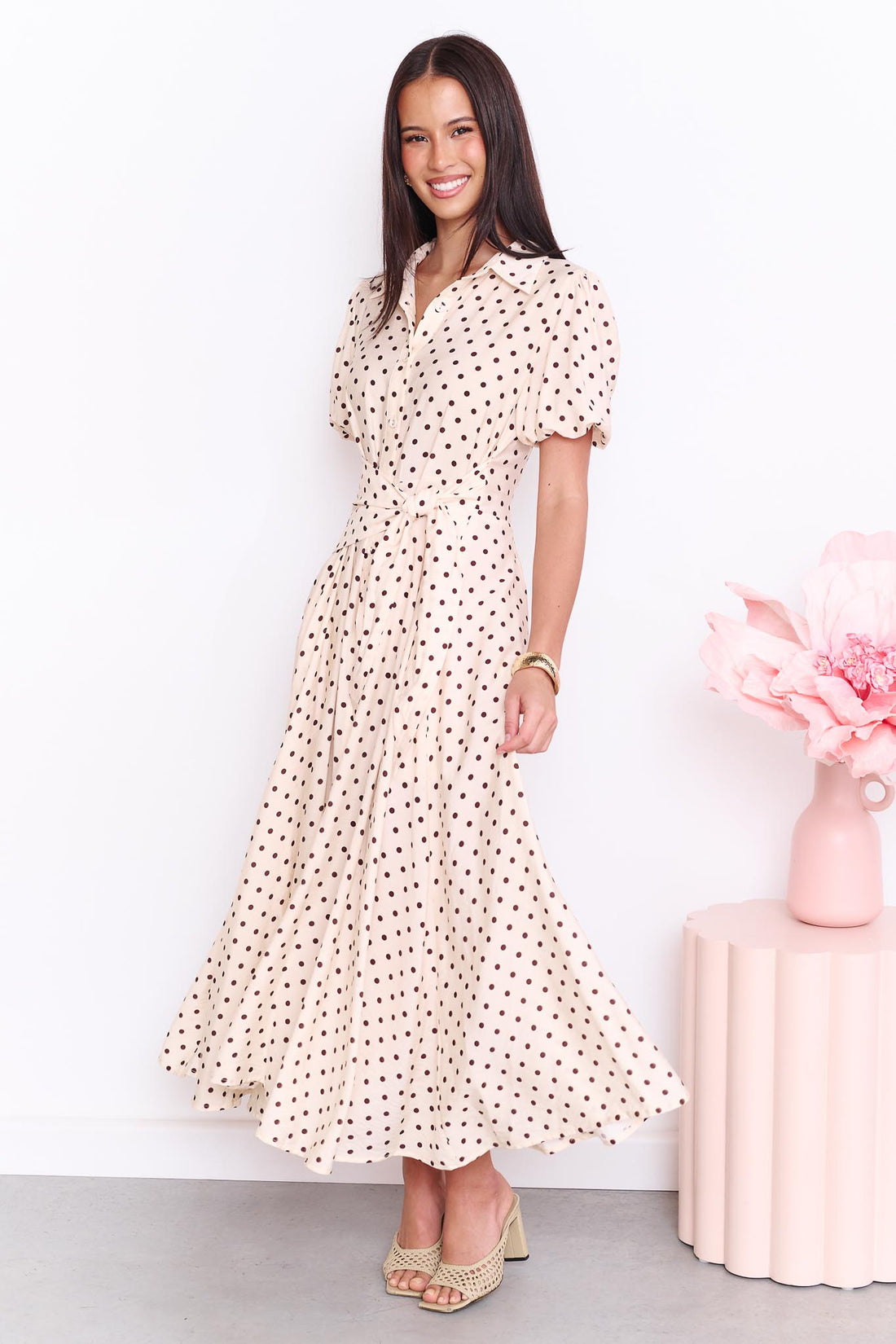 A model wearing the minnie + MAXXIE Viella Midi Dress in buttermilk with brown polka dots and a side waist tie, standing in front of a white wall.