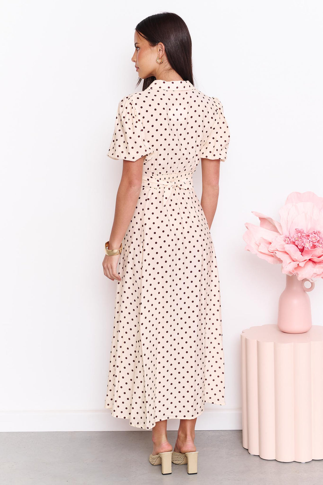 Back view of a model wearing the minnie + MAXXIE Viella Midi Dress in buttermilk with brown polka dots and a back waist tie, standing in front of a white wall.