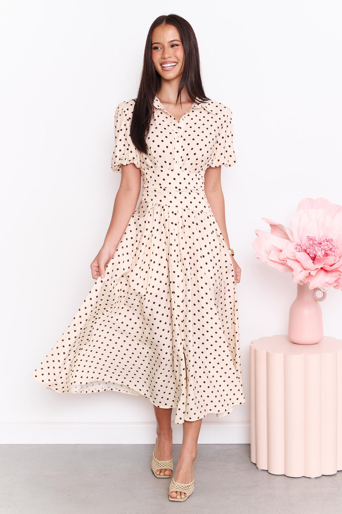 A model wearing the minnie + MAXXIE Viella Midi Dress in buttermilk with brown polka dots and a crossover waist, standing in front of a white wall.