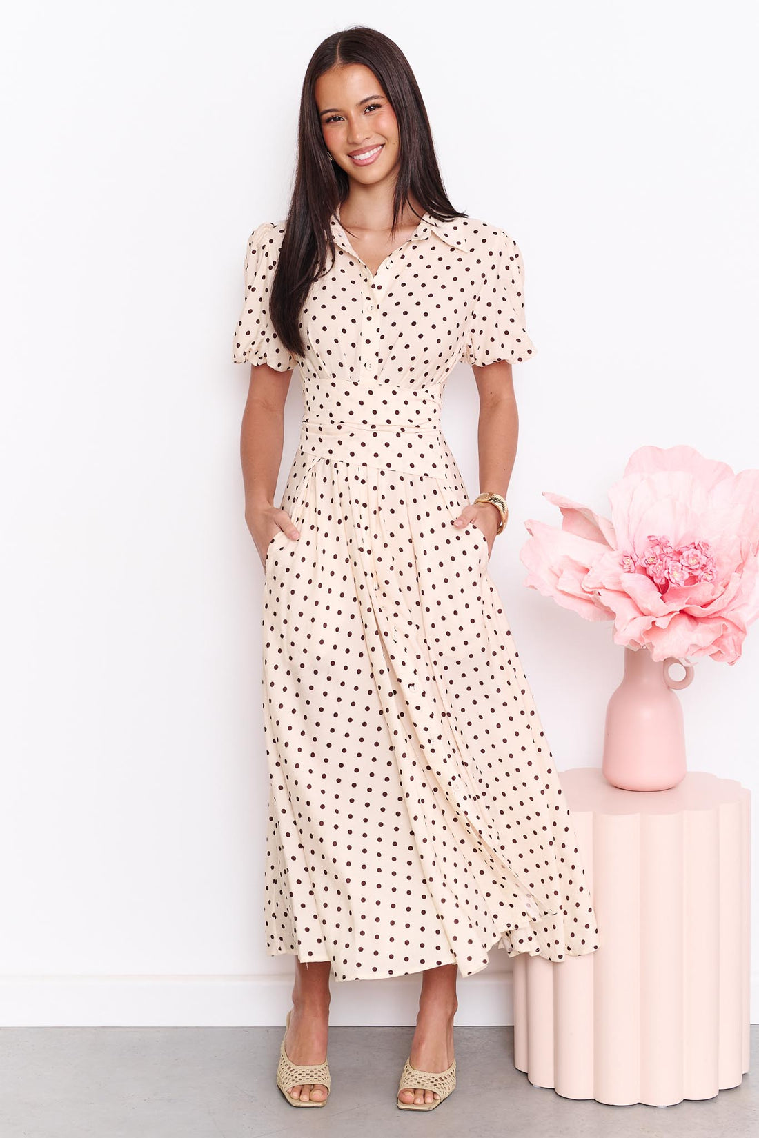 A model wearing the minnie + MAXXIE Viella Midi Dress in buttermilk with brown polka dots and a crossover waist, standing in front of a white wall.