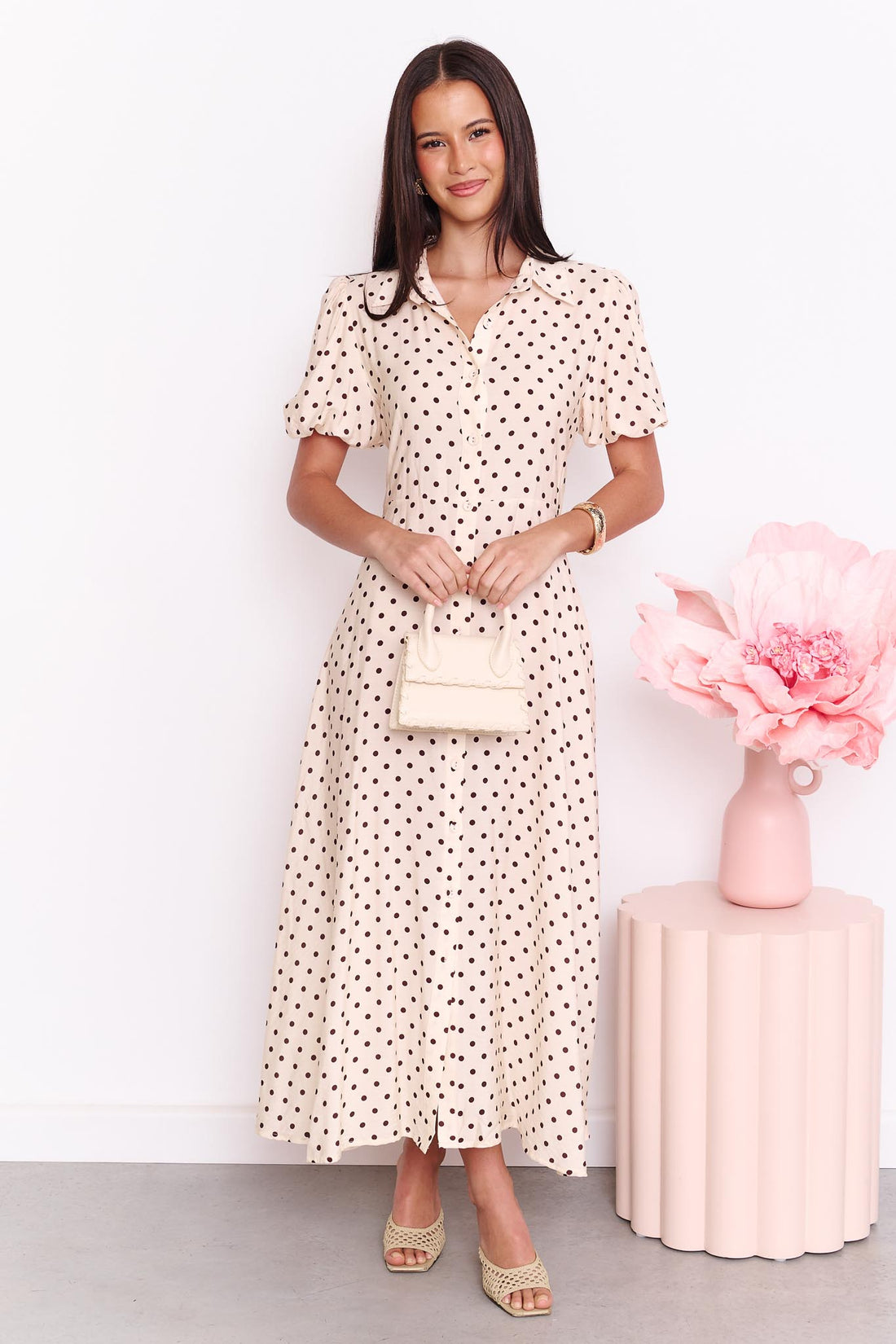A model wearing the minnie + MAXXIE Viella Midi Dress in buttermilk with brown polka dots and button-through front, standing in front of a white wall.