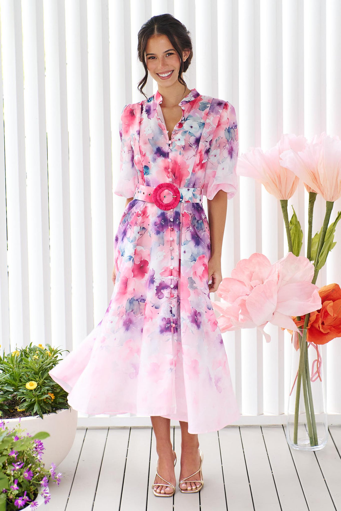 A woman wearing the minnie + MAXXIE Willow Bloom midi dress with puff sleeves and a pink and purple watercolour print with pink raffia belt, standing in front of a white panelled wall.