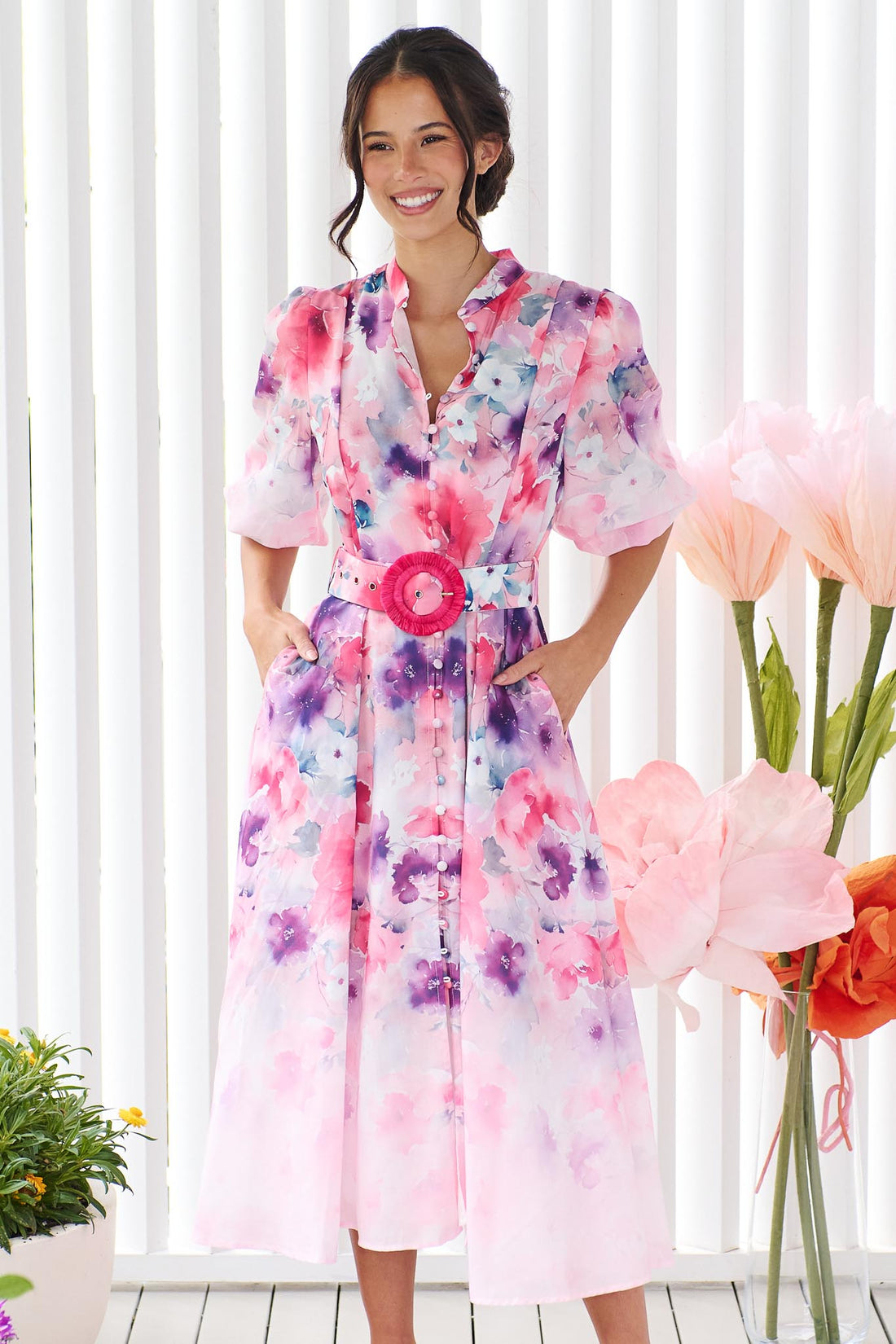 A woman wearing the minnie + MAXXIE Willow Bloom midi dress with puff sleeves and a pink and purple watercolour print with pink raffia belt, standing in front of a white panelled wall.