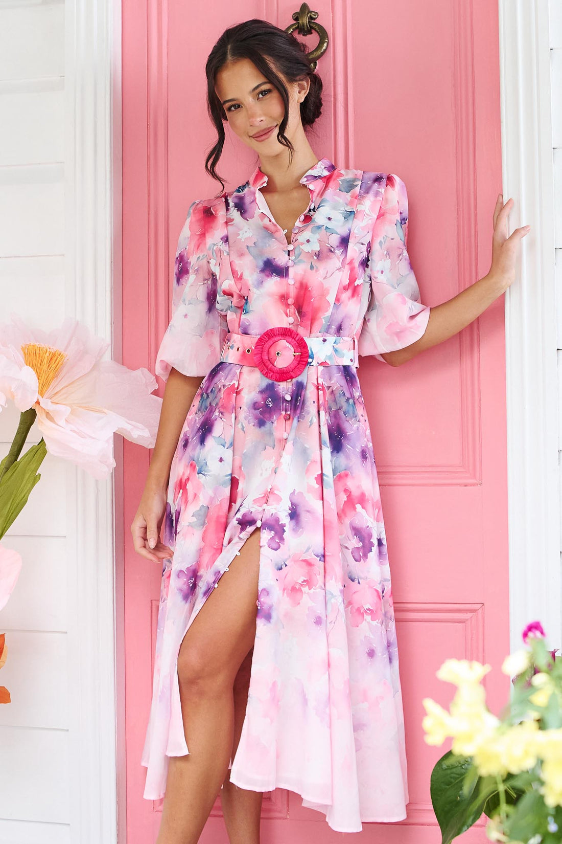 A woman wearing the minnie + MAXXIE Willow Bloom midi dress with puff sleeves and a pink and purple watercolour print with pink raffia belt, standing in front of a pink door.