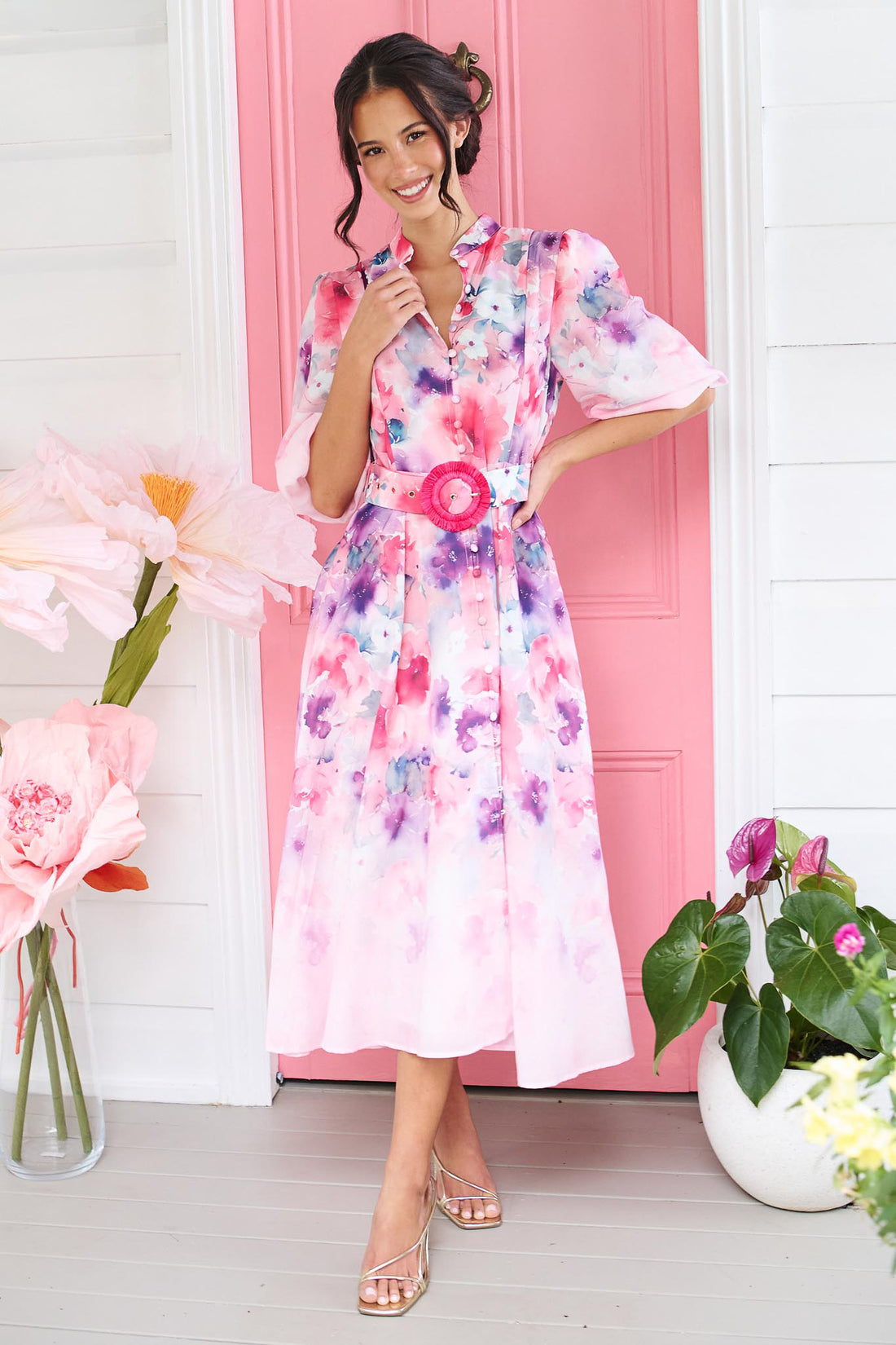 A woman wearing the minnie + MAXXIE Willow Bloom midi dress with puff sleeves and a pink and purple watercolour print with pink raffia belt, standing in front of a pink door.
