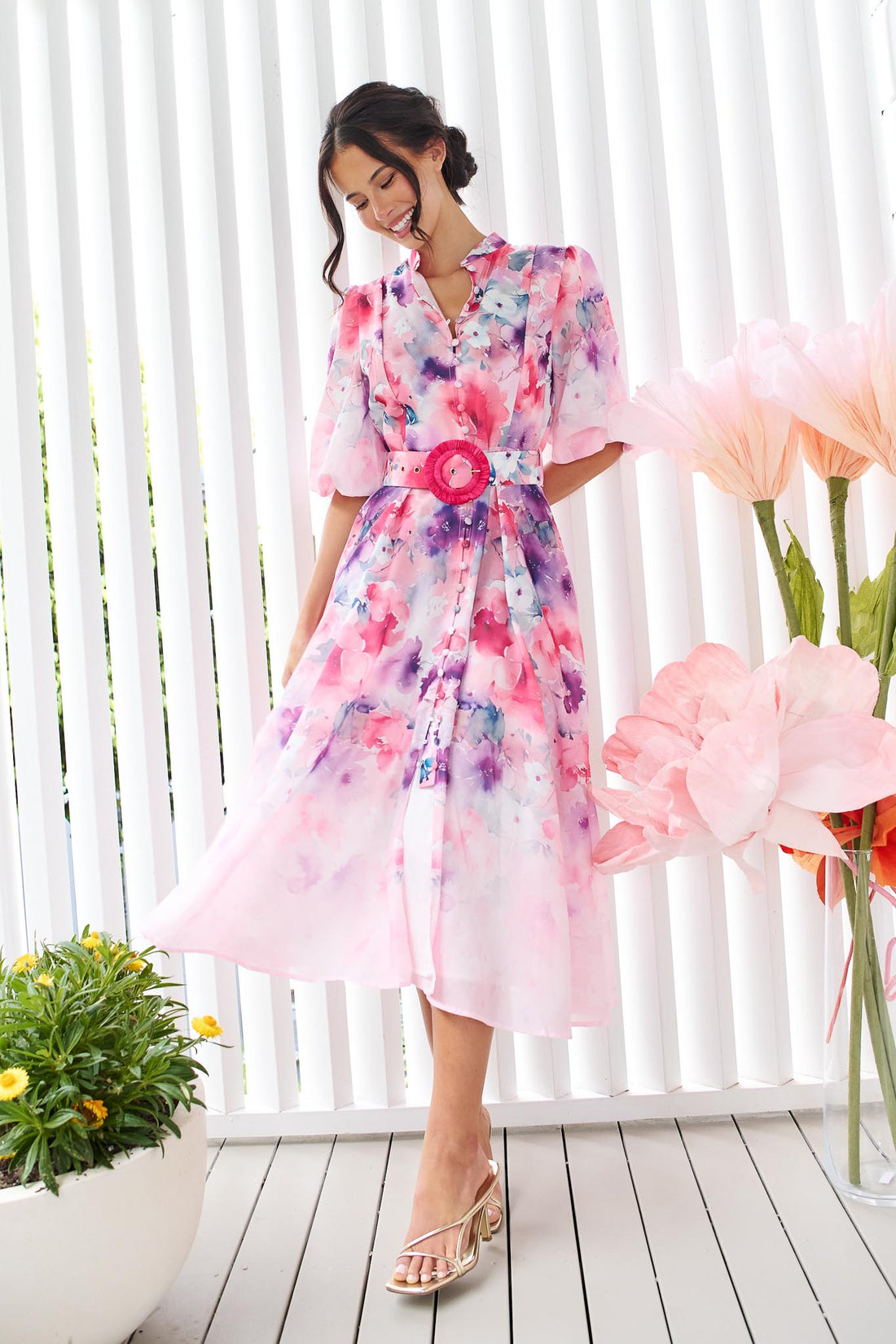 A woman wearing the minnie + MAXXIE Willow Bloom midi dress with puff sleeves and a pink and purple watercolour print with pink raffia belt, standing in front of a white panelled wall.