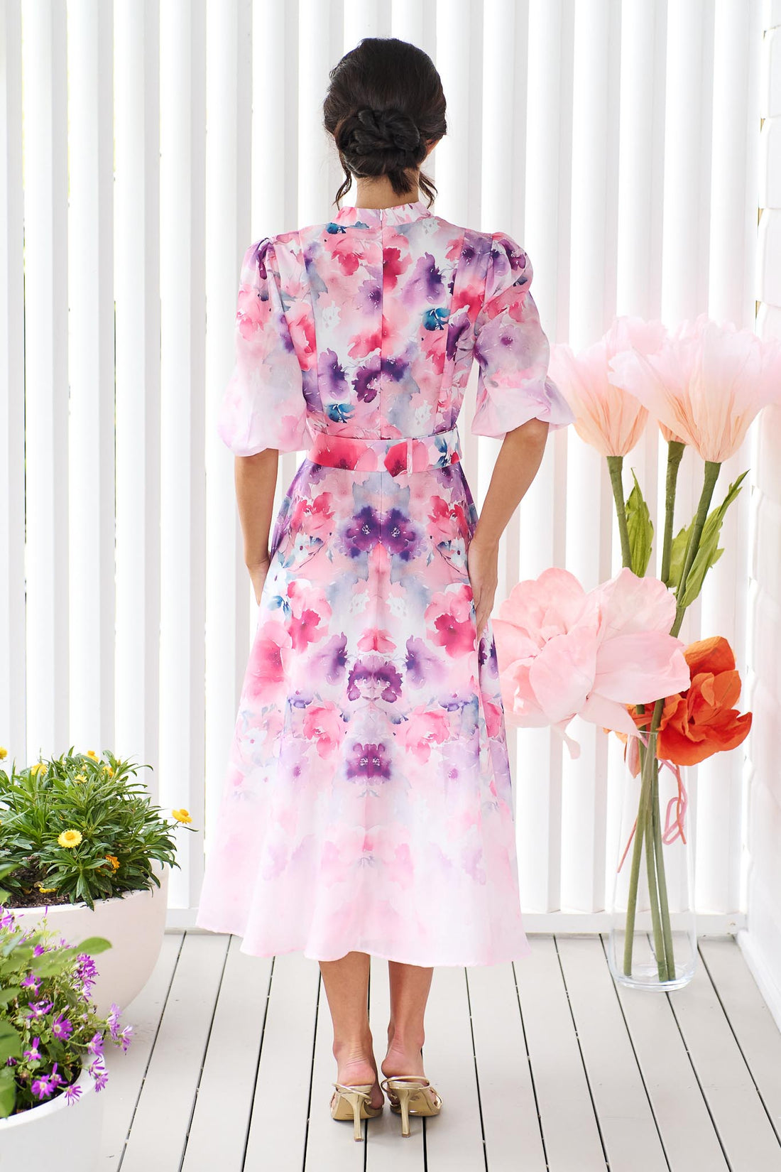 Back view of a woman wearing the minnie + MAXXIE Willow Bloom midi dress with puff sleeves and a pink and purple watercolour print with pink raffia belt, standing in front of a white panelled wall.