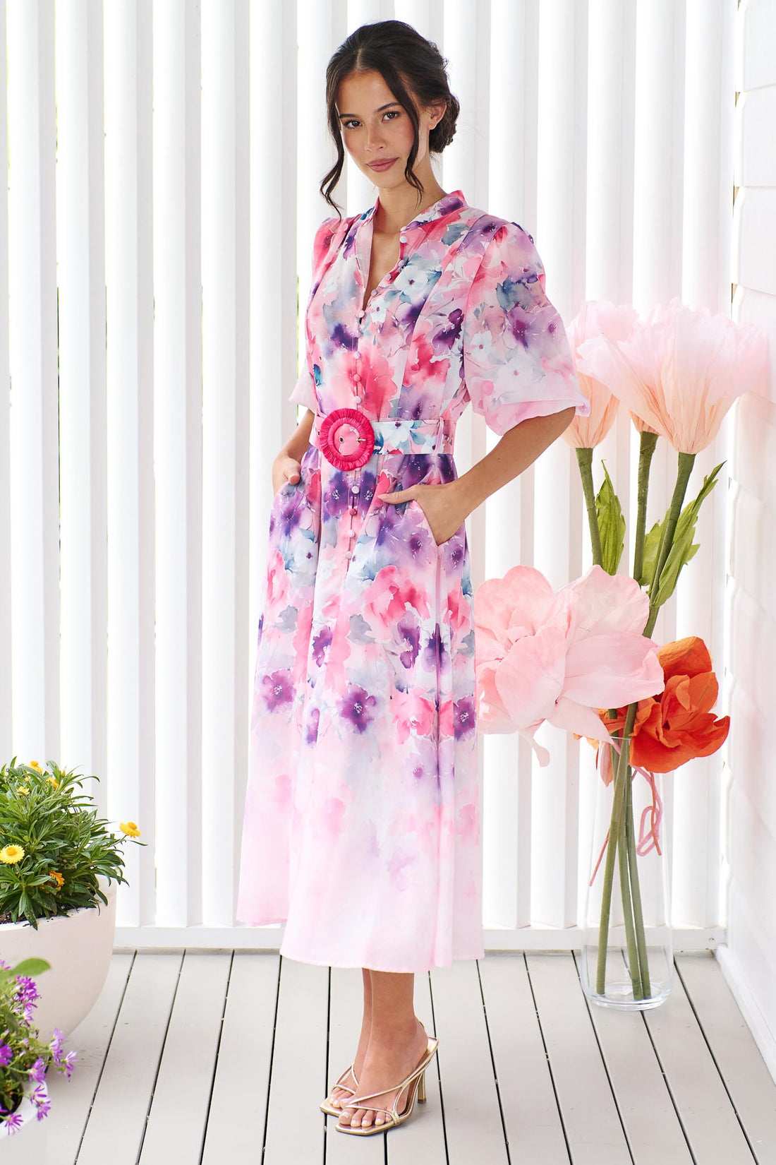 A woman wearing the minnie + MAXXIE Willow Bloom midi dress with puff sleeves and a pink and purple watercolour print with pink raffia belt, standing in front of a white panelled wall.