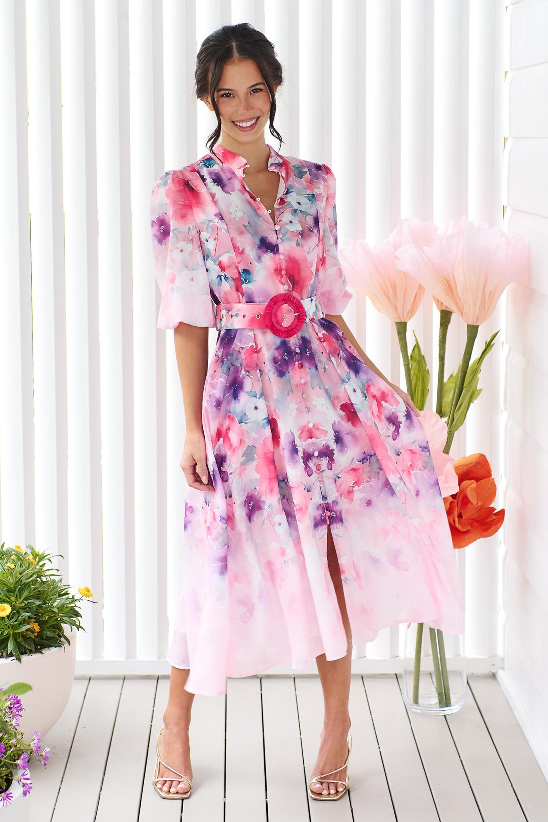 A woman wearing the minnie + MAXXIE Willow Bloom midi dress with puff sleeves and a pink and purple watercolour print with pink raffia belt, standing in front of a white panelled wall.